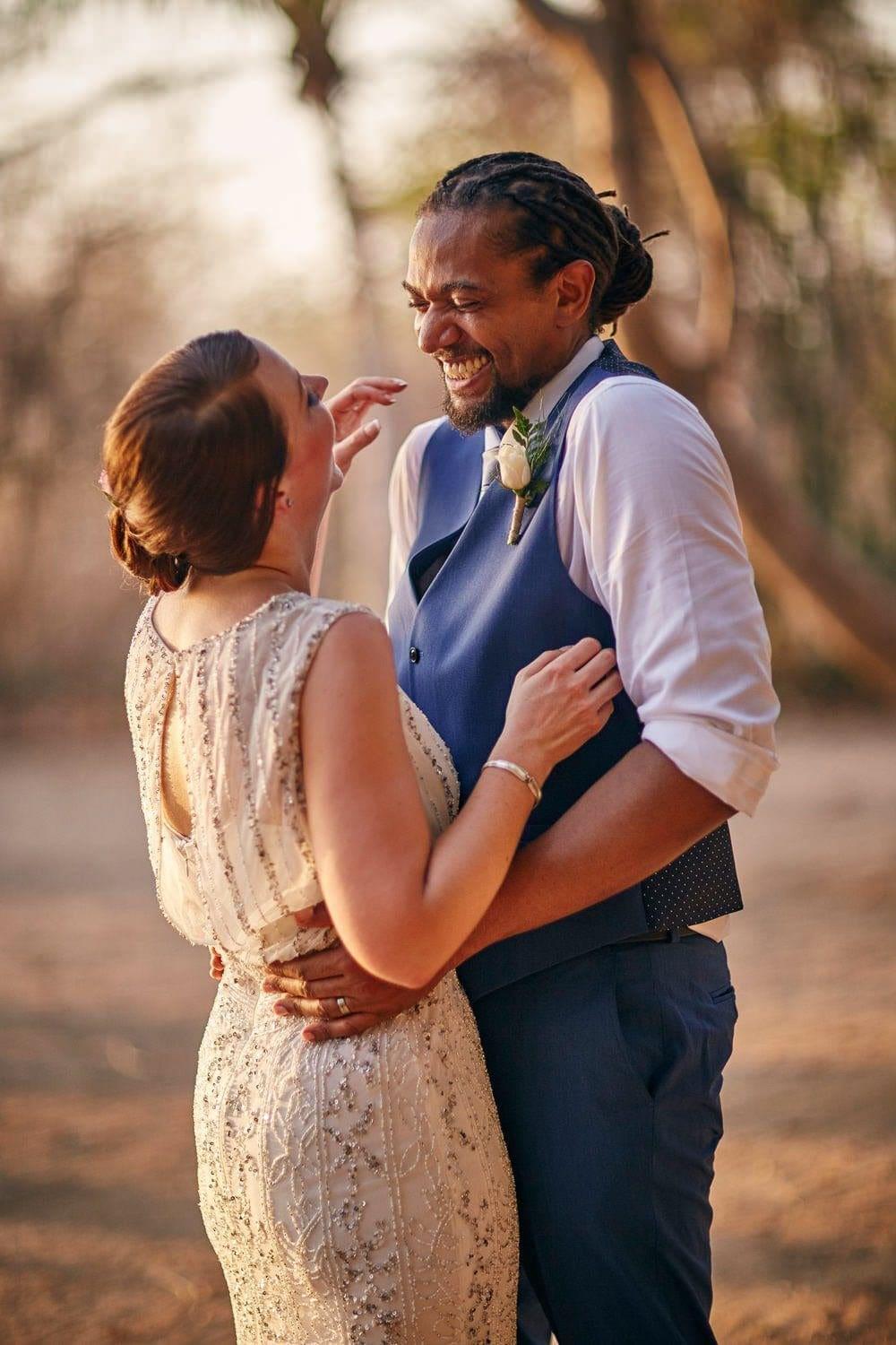 A candid golden hour portrait of a bride and groom laughing together during their Costa Rica destination wedding. The bride wears a beaded vintage style gown and the groom is dressed in a blue vest and white shirt, captured in a natural outdoor setting by a Costa Rica wedding photographer known for emotional, documentary style wedding photography.