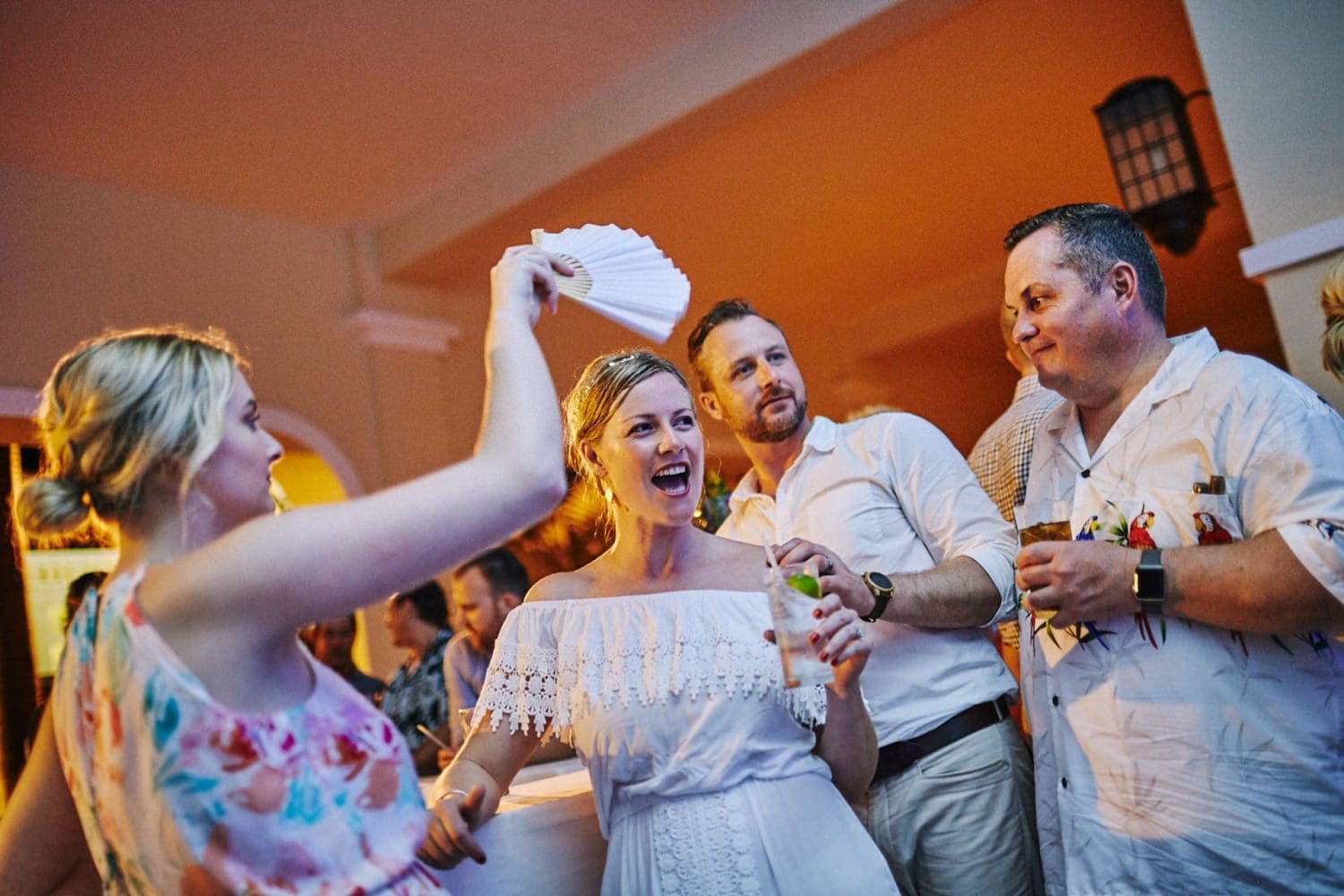 A guest fanning the laughing bride with a paper fan while friends stand around holding drinks at a wedding reception