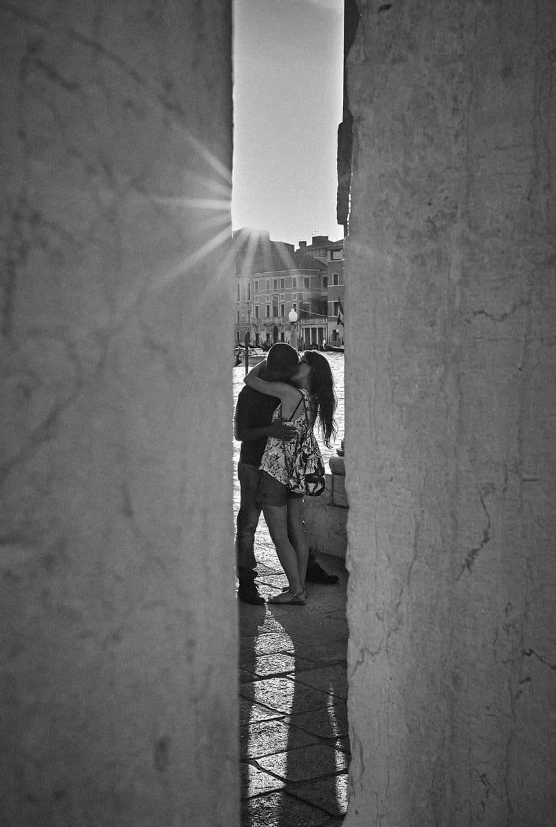 A couple kissing beside a Venetian canal, framed by a narrow gap between two walls with the sun behind them