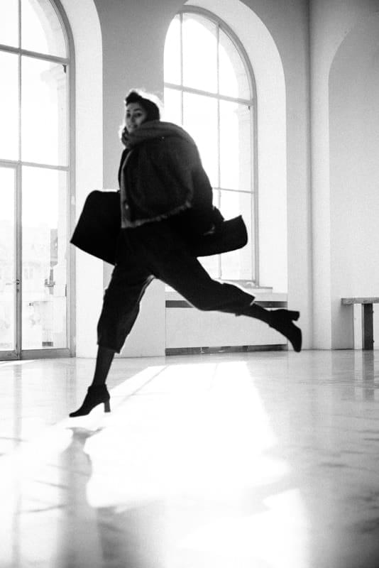 A black and white portrait of a woman in a dark coat and heels leaping across a bright room with tall arched windows