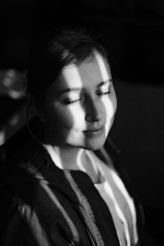 A black and white portrait of a young woman with her eyes closed, stripes of light falling across her face